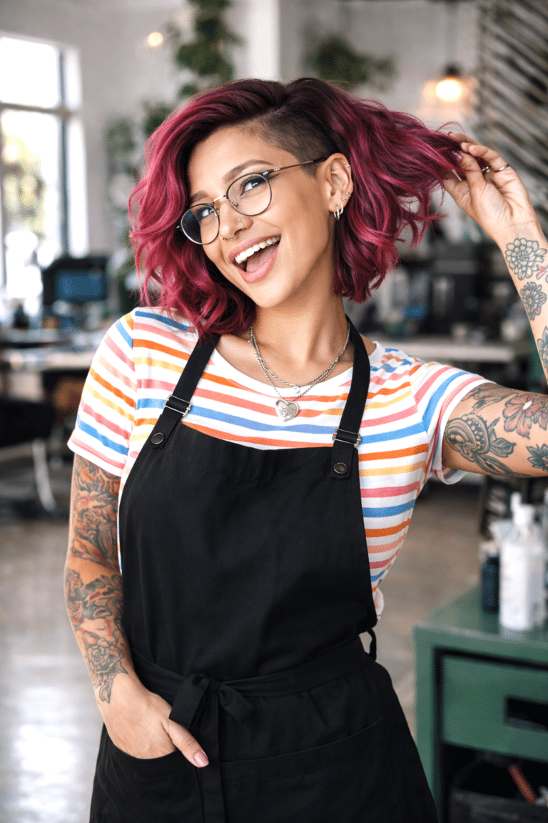 Smiling person with pink hair, glasses, and tattoos wears a striped shirt and black apron, standing in a modern, bright salon.