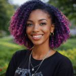 A woman with short curly purple hair, wearing hoop earrings, necklaces, and a black shirt, smiles outdoors in a park setting.