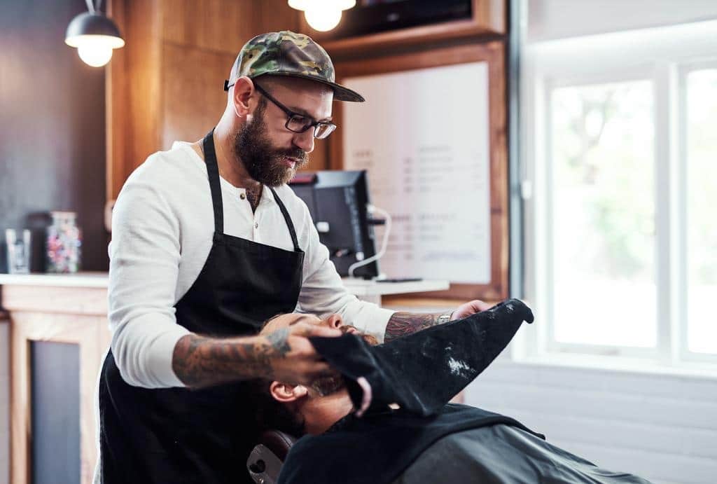 A barber wearing an apron and cap places a hot towel on a client's face in a barbershop.