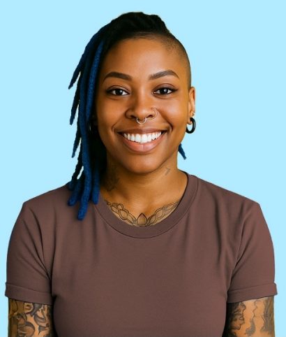 A person with blue-tipped locs, tattoos, and facial piercings smiles at the camera while wearing a brown t-shirt against a light blue background.